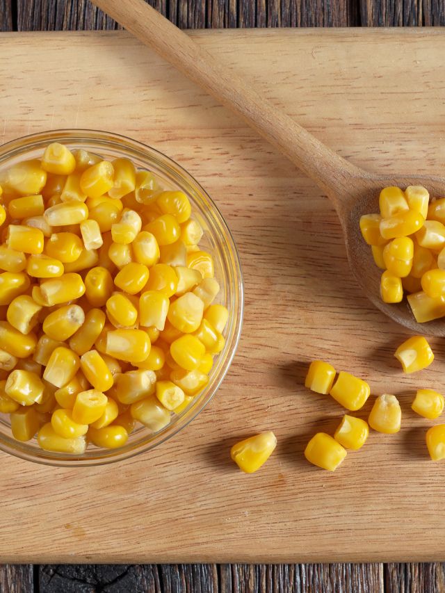 a bowl of corn with a wooden spoon of corn on a wooden cutting board