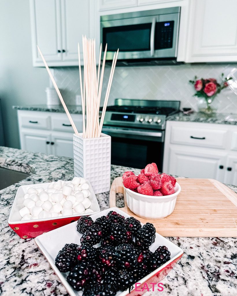 ingredients for american fruit flag featuring a bowl of blackberries, a bowl of raspberries, mini marshmallows and skewers