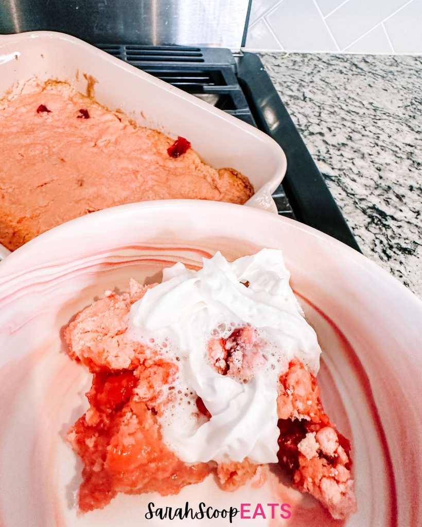 3 ingredient strawberry dump cake featuring cake with whipped cream in a bowl by a pan of strawberry cake