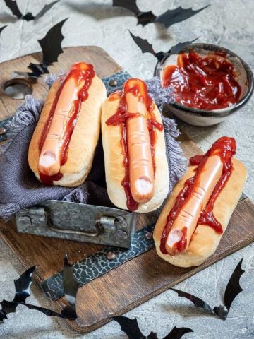 Three hot dogs with ketchup and bats on a wooden cutting board.