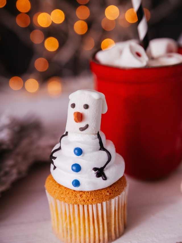 A cupcake with a snowman on top and a cup of hot cocoa.