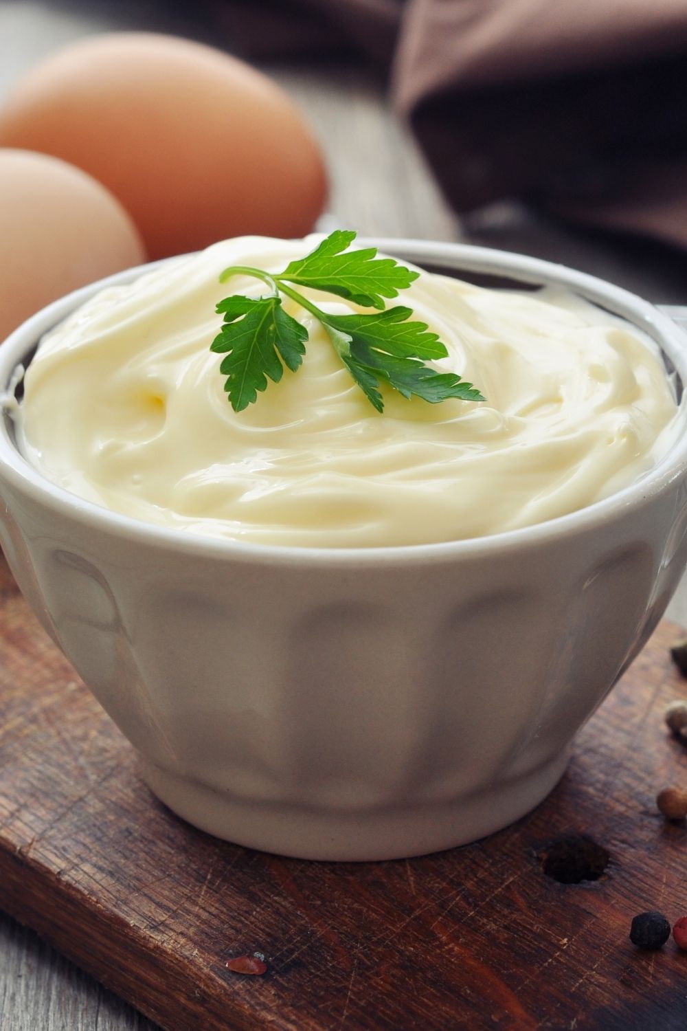 A bowl of creamy mayonnaise topped with a parsley leaf, surrounded by eggs and peppercorns on a wooden board.