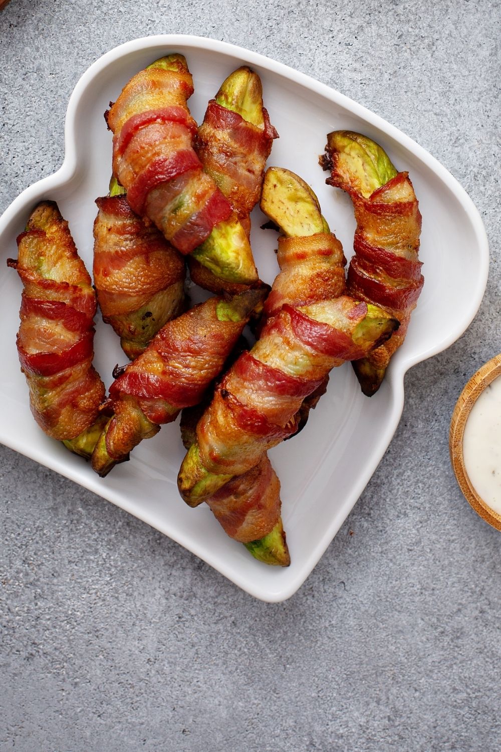 A white, heart-shaped plate holds bacon-wrapped avocado slices, arranged neatly—a perfect addition to your keto snack ideas. The background is a gray, textured surface with a small bowl of dipping sauce partially visible to the right.