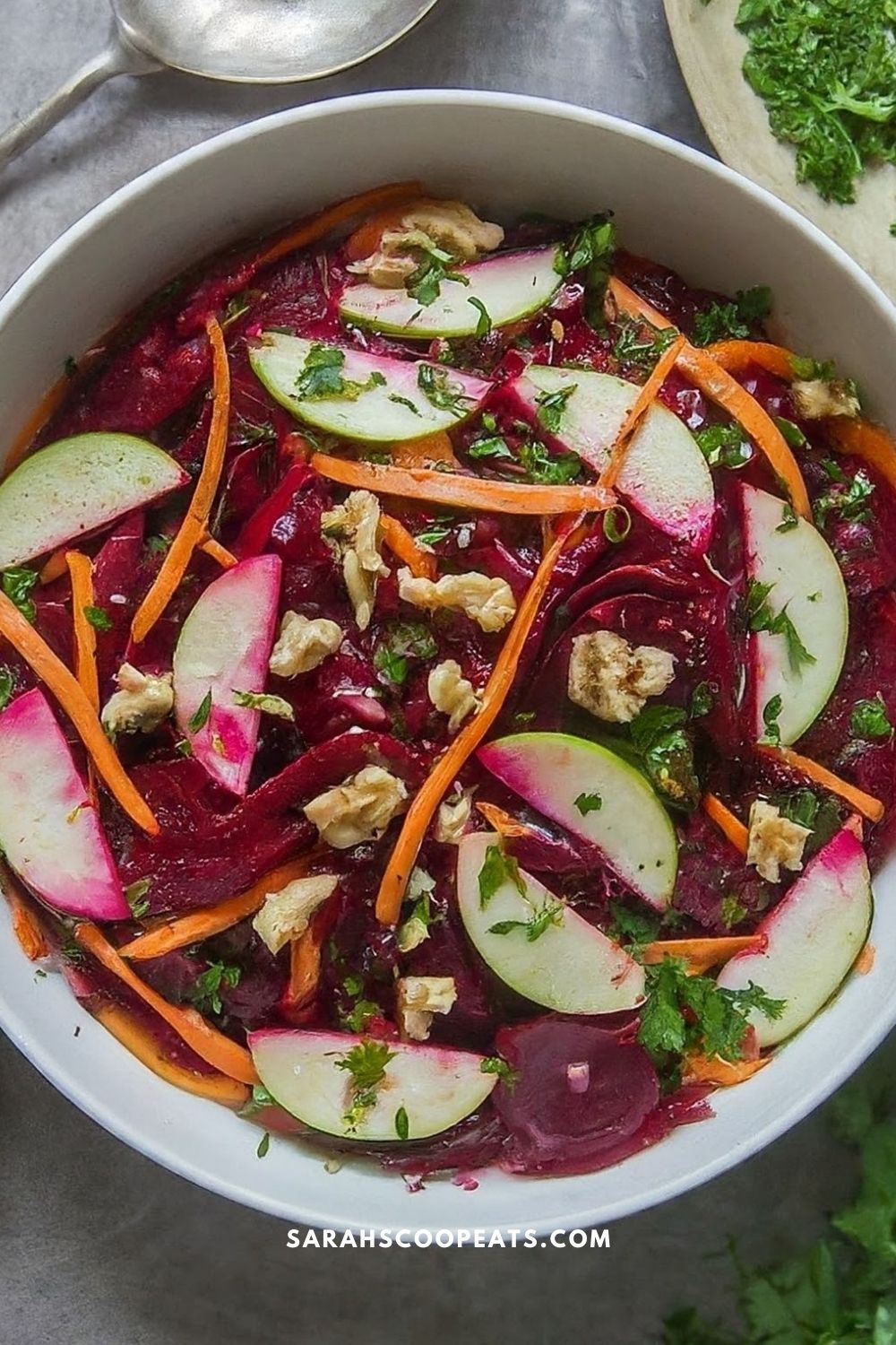 A bowl of beet salad with slices of radish, apple, carrot strips, chopped walnuts, and herbs, set on a table. Text at the bottom reads, "SARAHSHOPEATS.COM.
