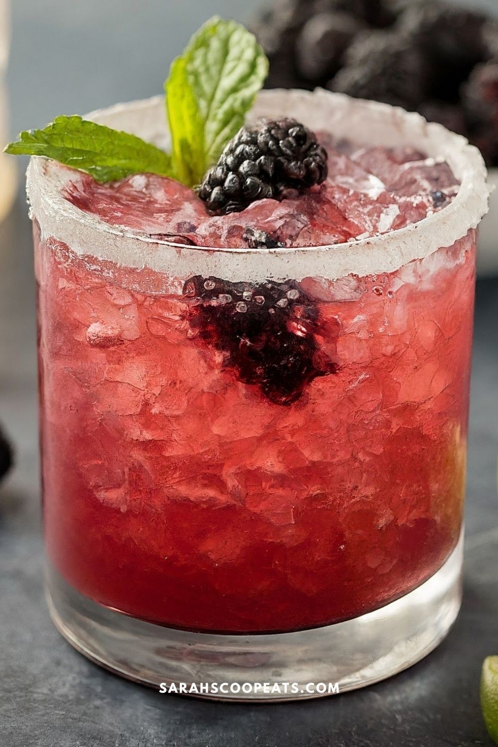 A glass of a refreshing berry cocktail, topped with a blackberry, ice, and a mint sprig. The drink is garnished with a sugar-rimmed rim.