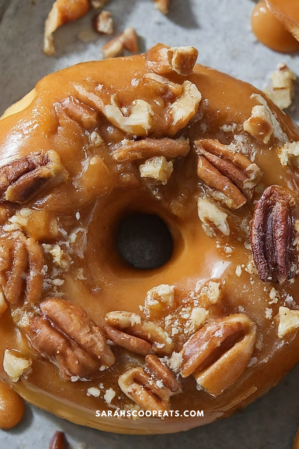 A close-up of a glazed doughnut topped with chopped pecans and bits of caramel, placed on a gray surface. Text on the image reads "SARAHSSCOPEATS.COM.