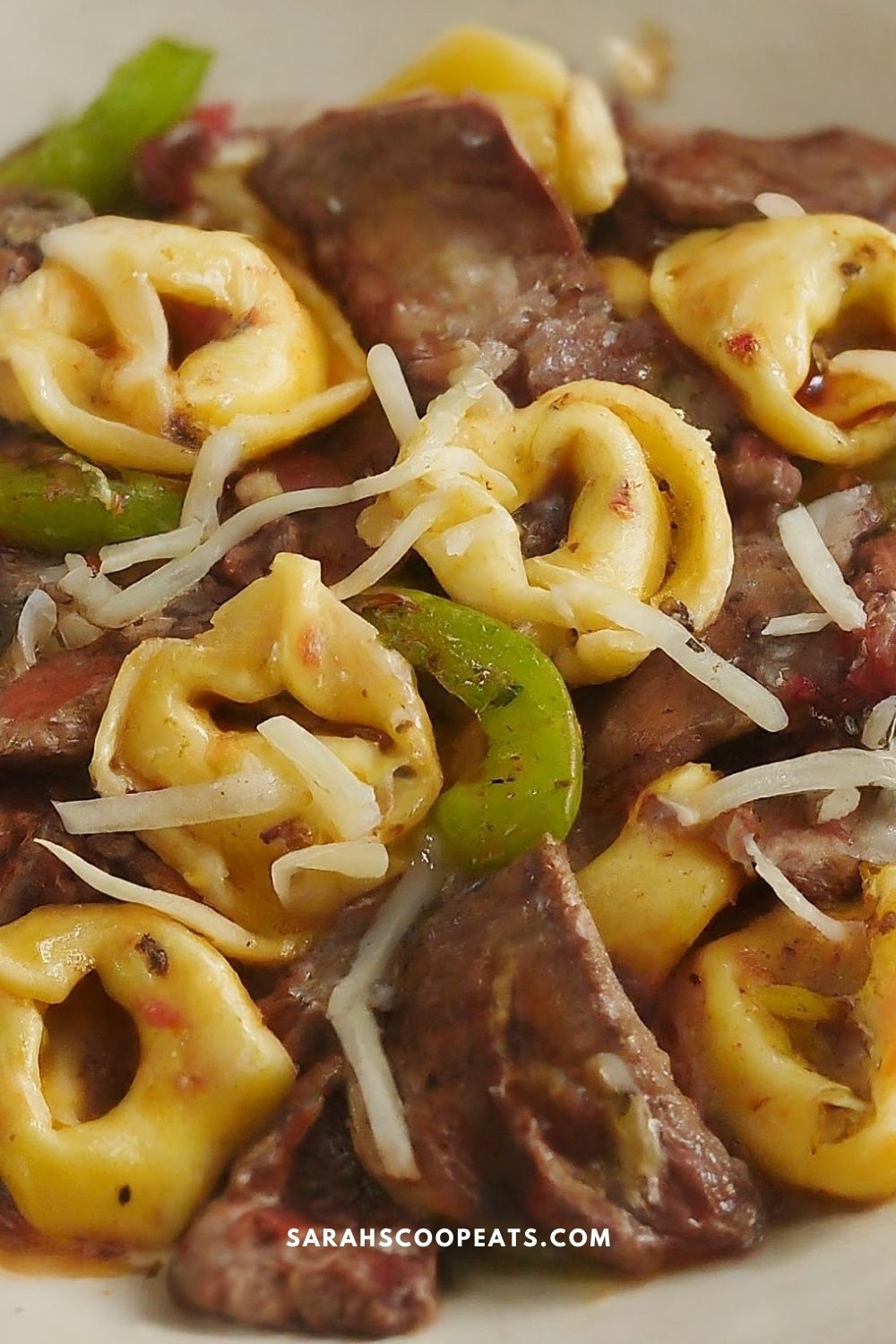 A close-up of a pasta dish with beef, tortellini, green bell peppers, and grated cheese.