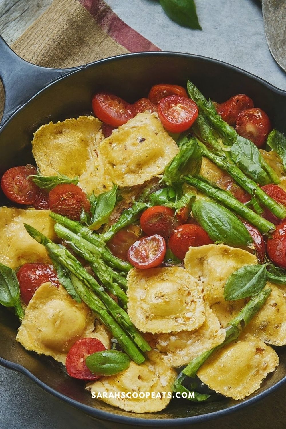 A skillet containing cooked cheese ravioli, cherry tomatoes, asparagus, and fresh basil leaves.