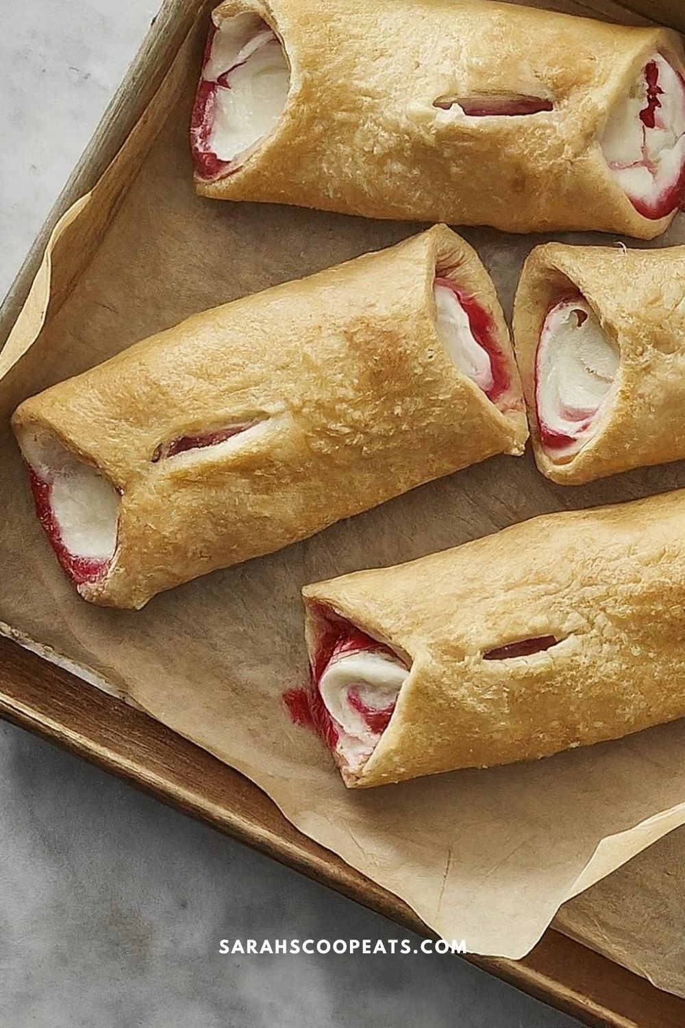 Four strawberry cream cheese danishes on a parchment-lined baking sheet. The pastries are golden brown with visible cream cheese and strawberry filling. The website sarahscoopeats.com is at the bottom.