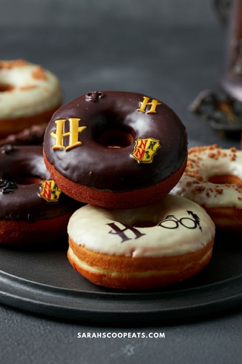 A plate of decorated donuts with chocolate and white frosting, featuring Harry Potter-themed symbols like H, Hogwarts crest, and golden snitch wings.