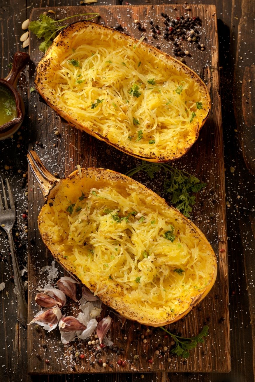 Two halved spaghetti squashes filled with cooked spaghetti-like strands, garnished with herbs, on a wooden cutting board, surrounded by garlic cloves, herbs, and seasoning.