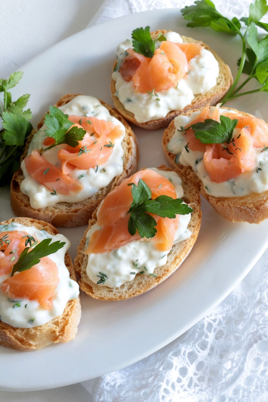 A plate of five bruschettas with smoked salmon, cream cheese, and herbs, garnished with parsley, on a white lace-trimmed cloth.