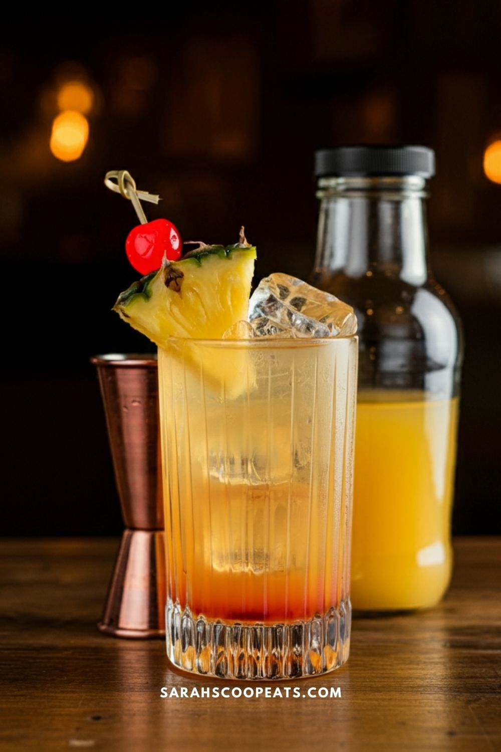 A cocktail garnished with a pineapple slice and cherry is served in a glass with ice, accompanied by a small bottle of orange juice and a jigger.