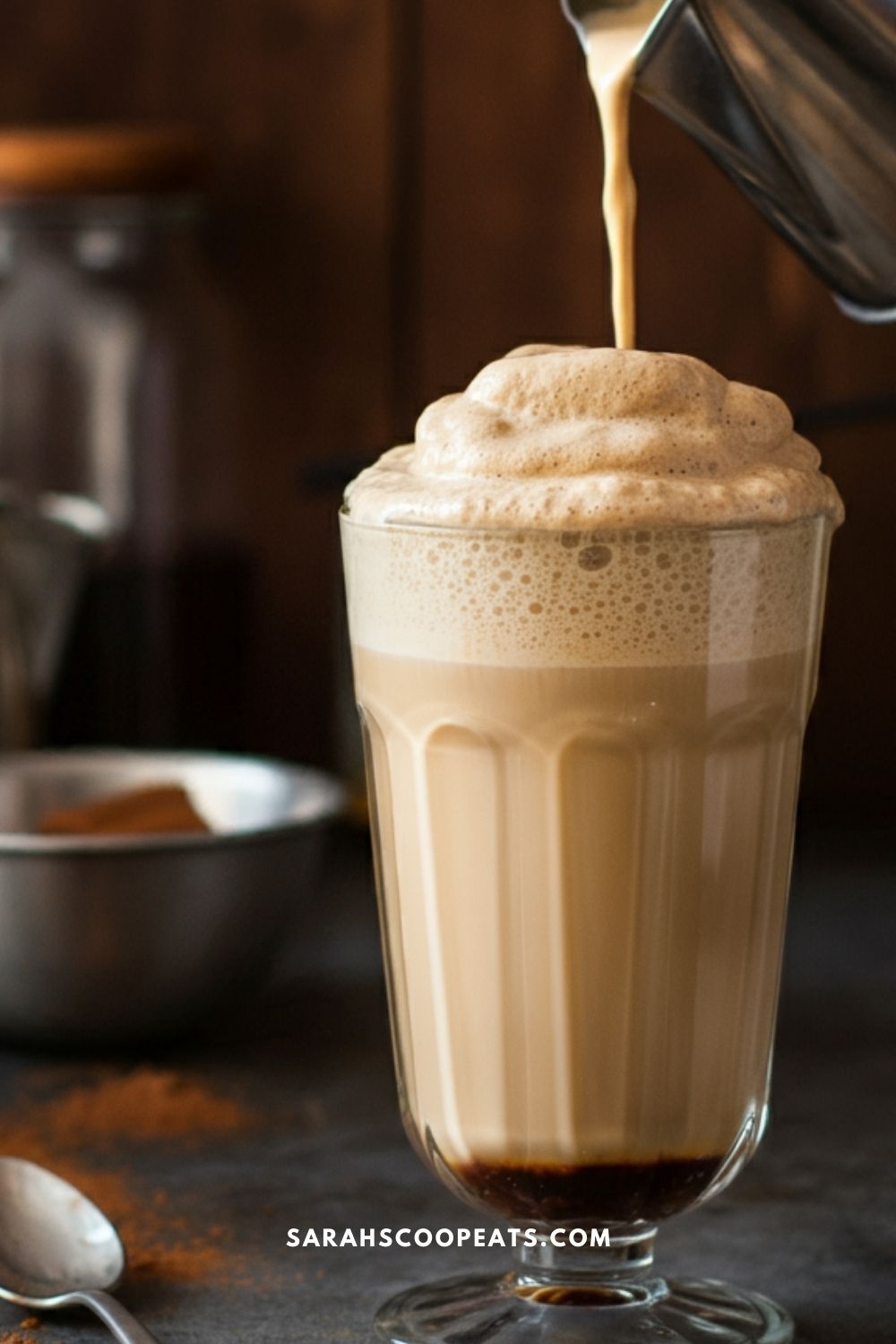 A glass filled with a frothy latte being poured from a metal container, with a spoon and jar in the background.