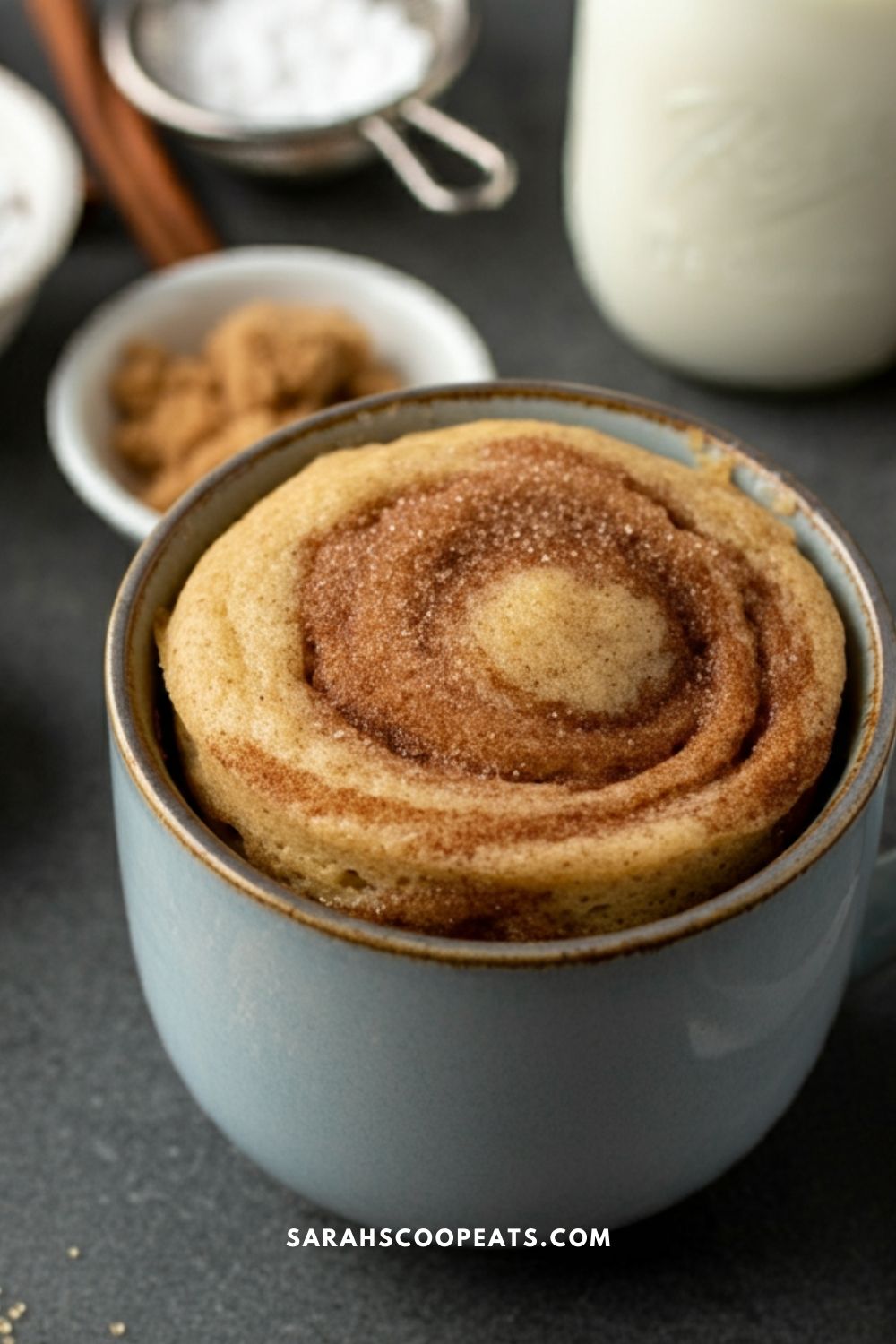 A cinnamon roll in a mug on a dark surface, with ingredients like brown sugar, salt, and a bottle of milk in the background.