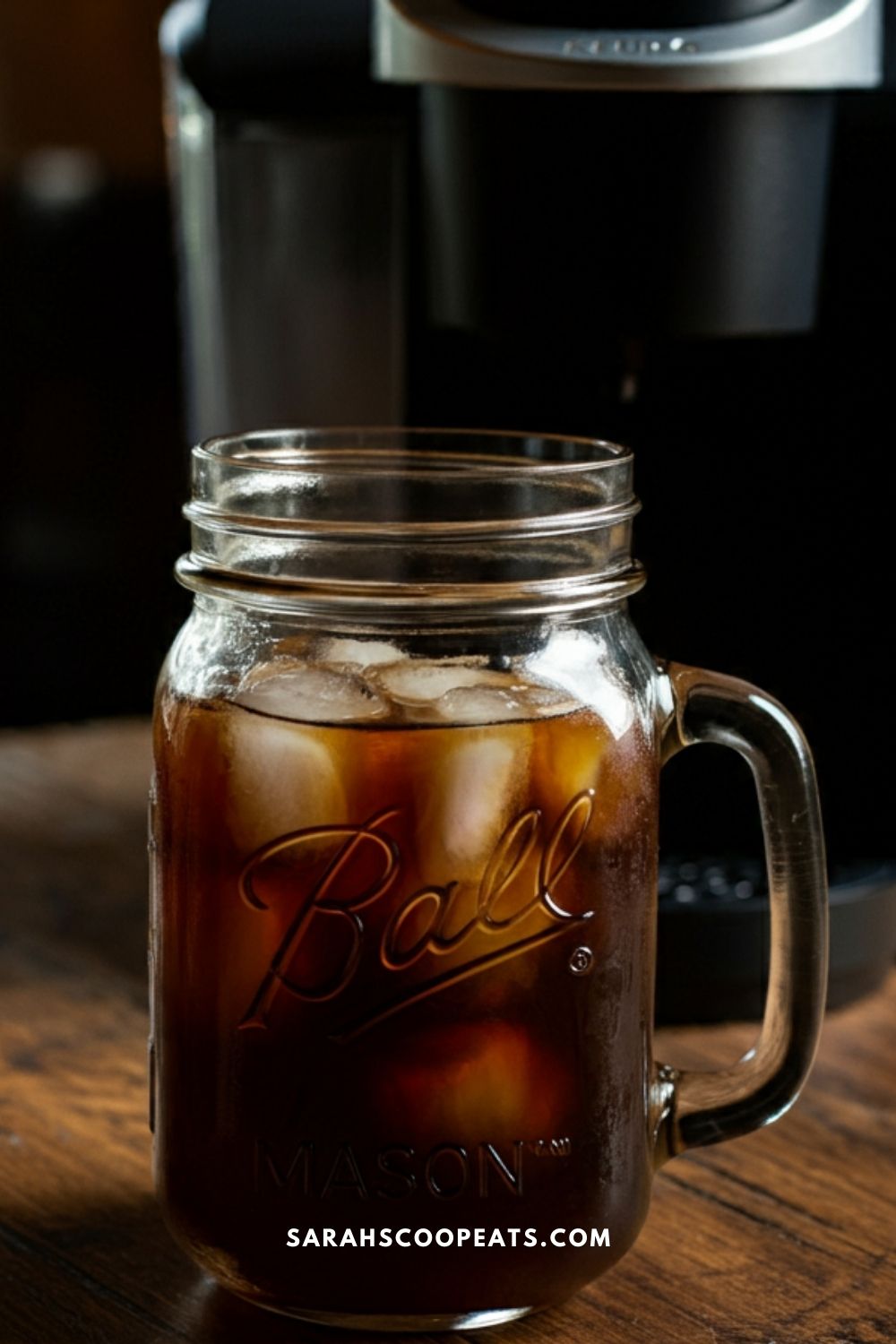 A mason jar filled with iced coffee sits on a wooden surface, in front of a coffee maker. The jar is embossed with the word "Ball.