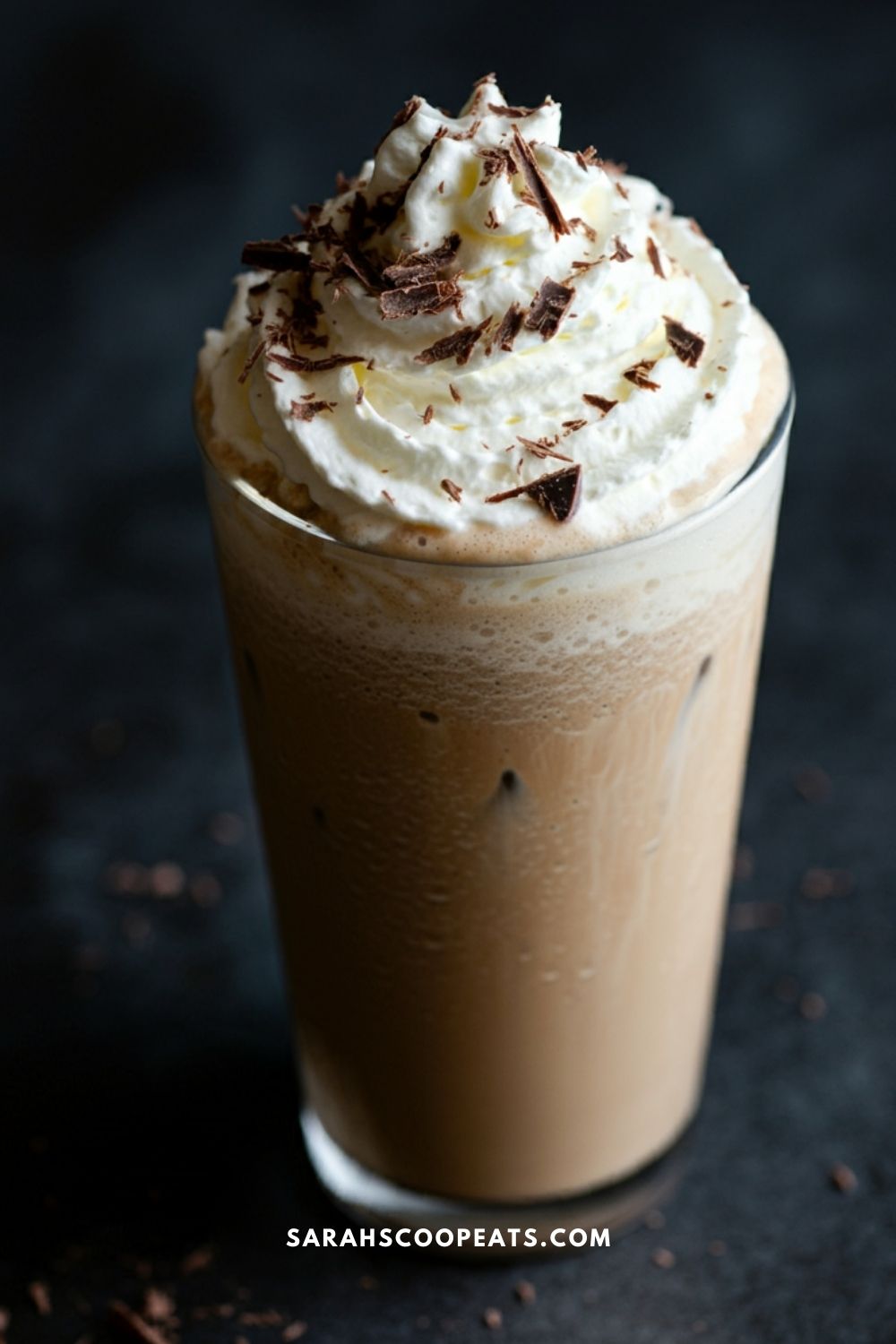 Tall glass of iced coffee topped with whipped cream and chocolate shavings on a dark background.