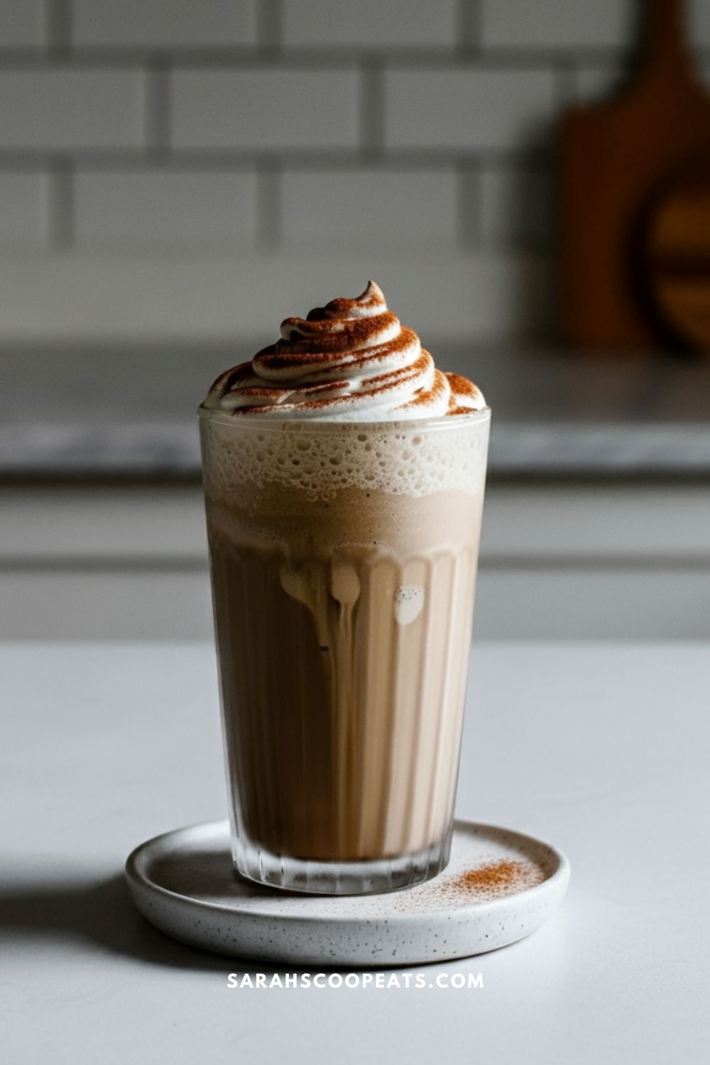 A frothy iced coffee frappuccino topped with whipped cream and a sprinkle of cocoa powder sits elegantly in a tall glass, placed on a small plate.