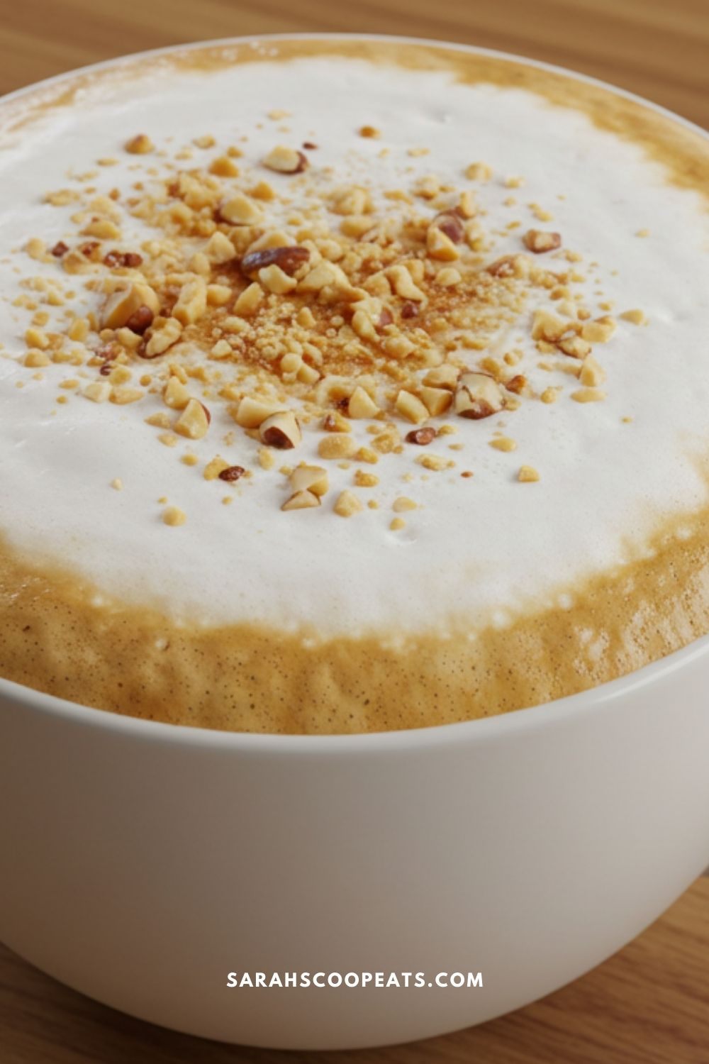 A cup of coffee topped with frothy milk and crushed nuts on a wooden table.