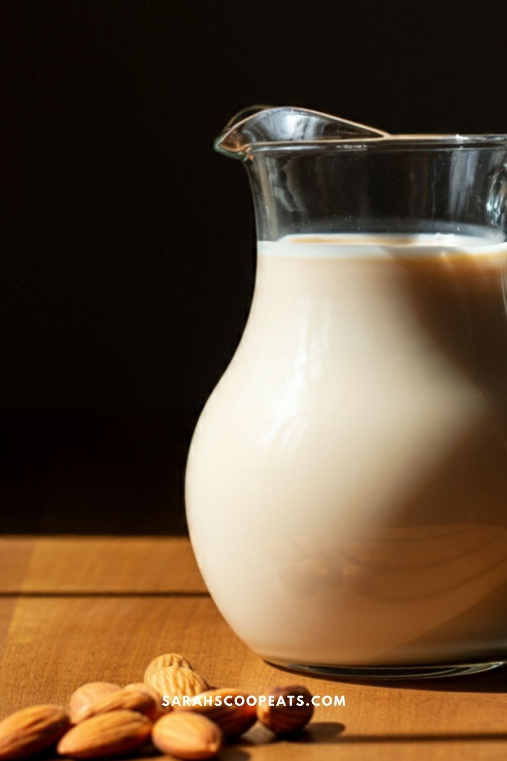 A glass pitcher filled with almond milk sits on a wooden surface, next to several almonds.
